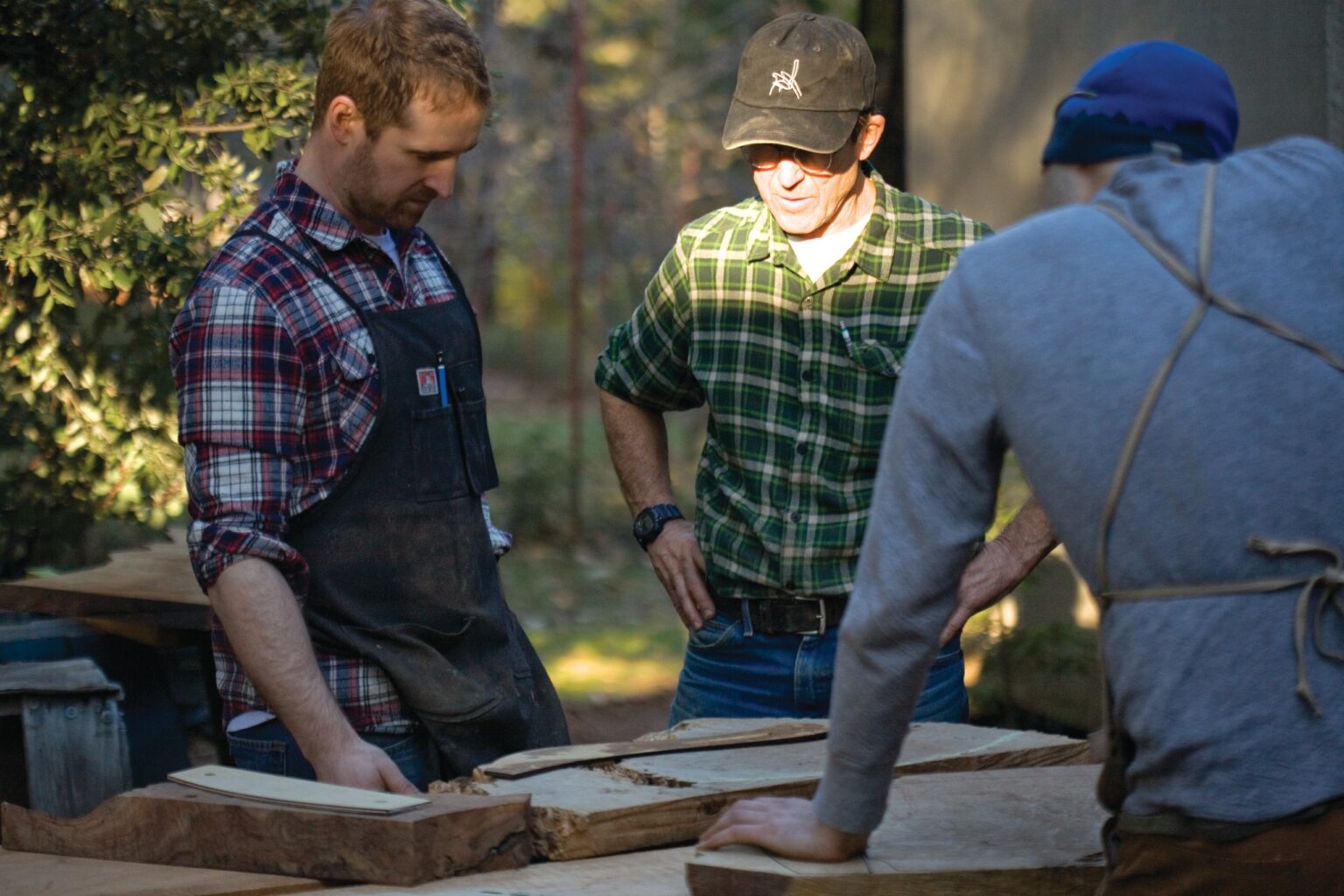 Three people examine a piece of wood outdoors.